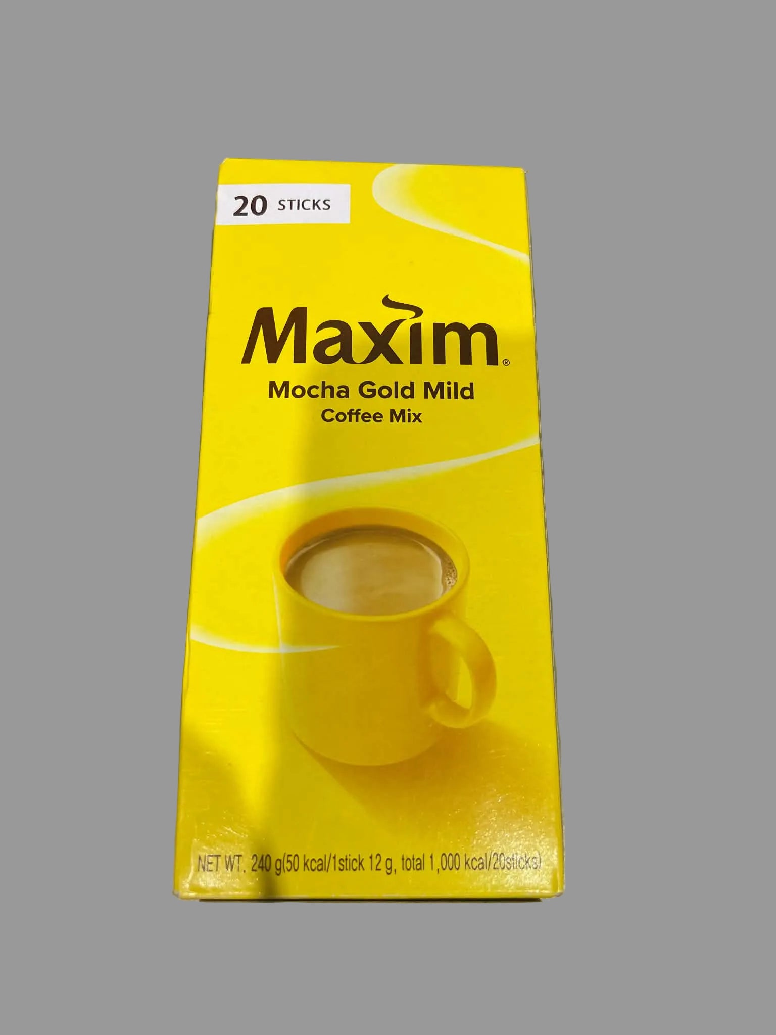 A yellow box of Maxim Mocha Gold Mild Coffee Mix, containing 20 sticks, placed on a gray surface.