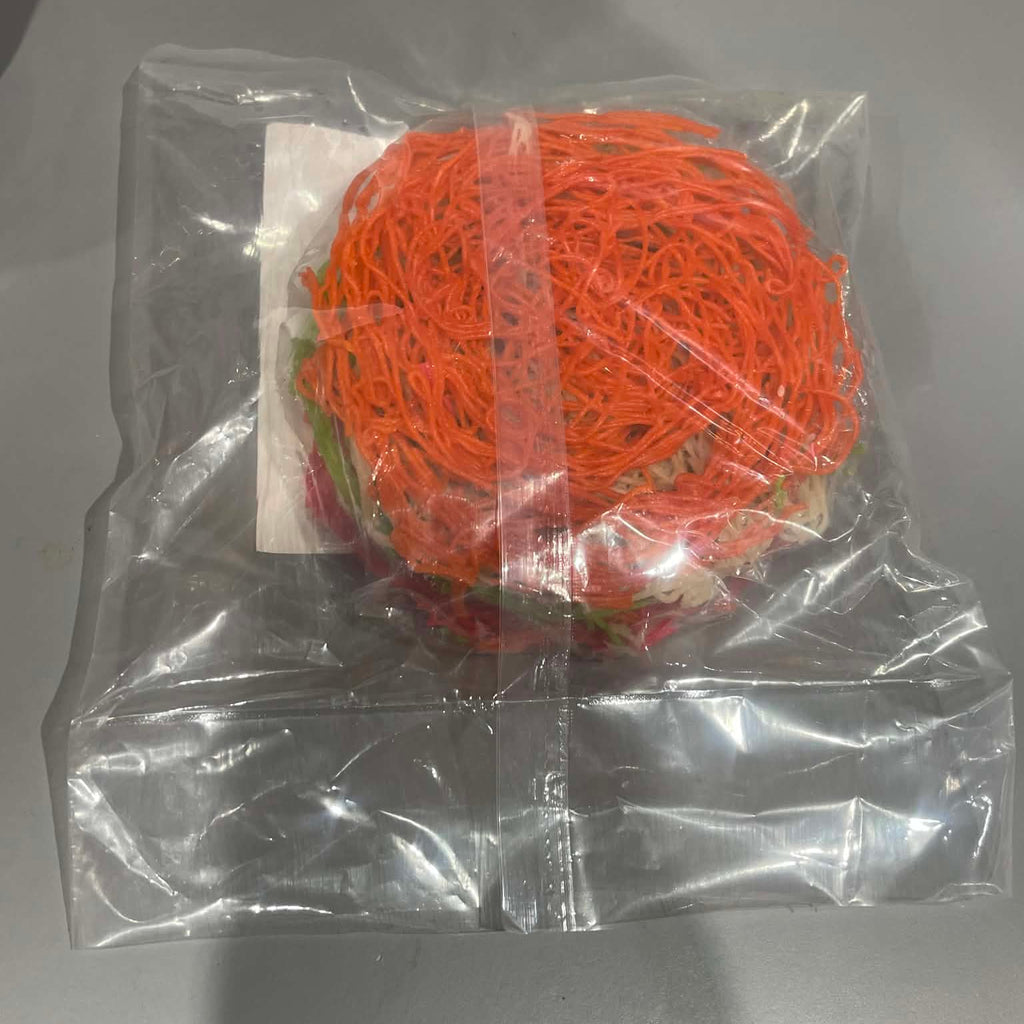 A round bundle of bright orange fuzzy strands, likely a decorative faux fur or pet toy, sealed inside a clear plastic bag with a white paper label, set against a plain gray background.