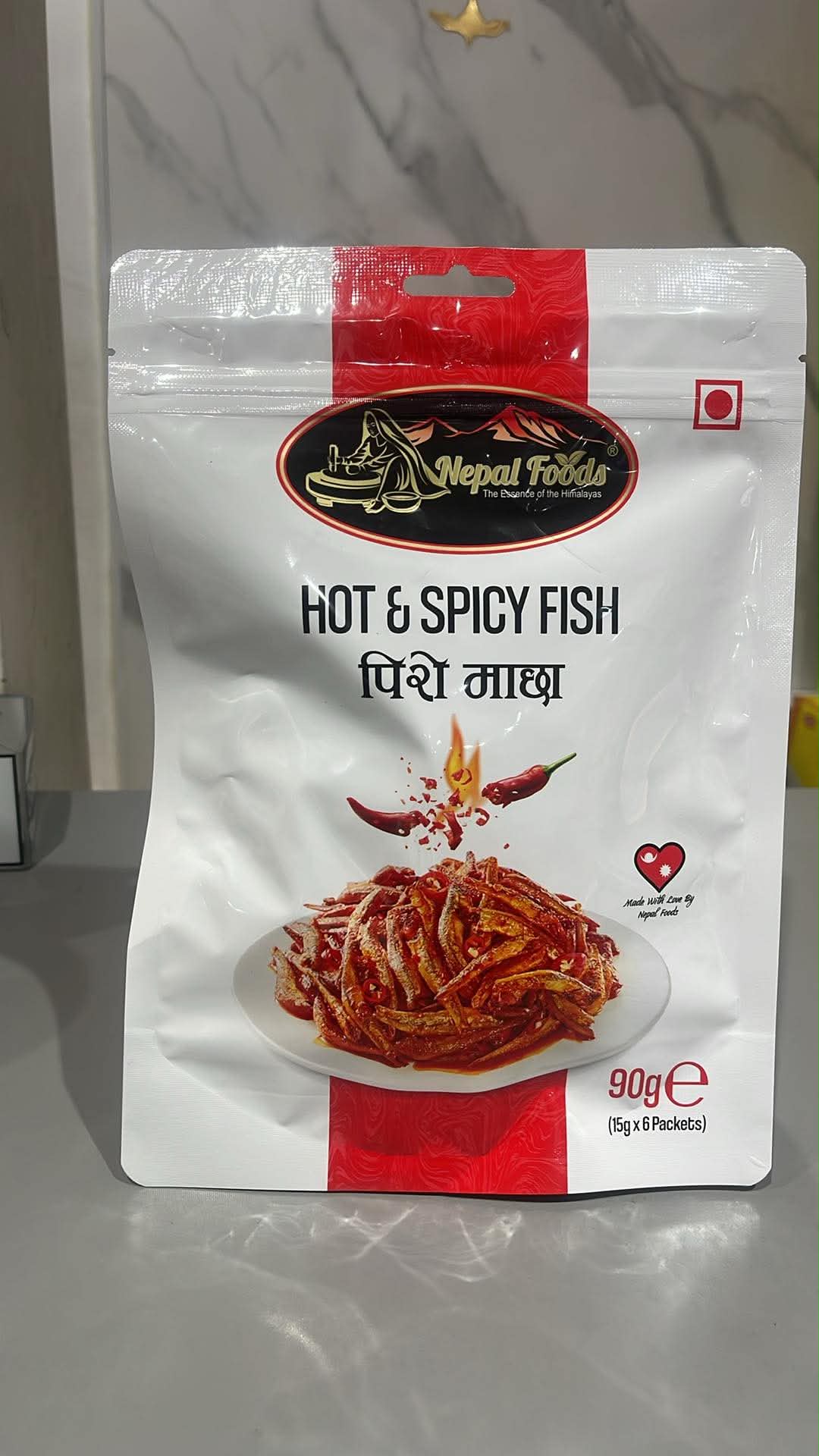 A 90g pouch of Nepal Fodds Hot & Spicy Fish (Pisni Maacha) in a white and red resealable stand-up bag, displayed on a gray countertop.