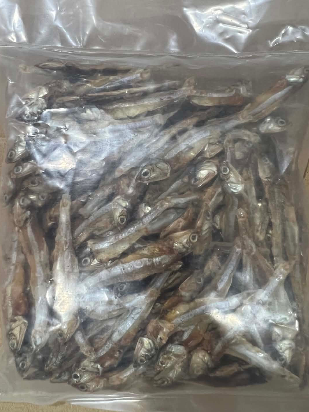 A clear plastic bag filled with dried small fish, photographed from above on a brown cardboard surface.