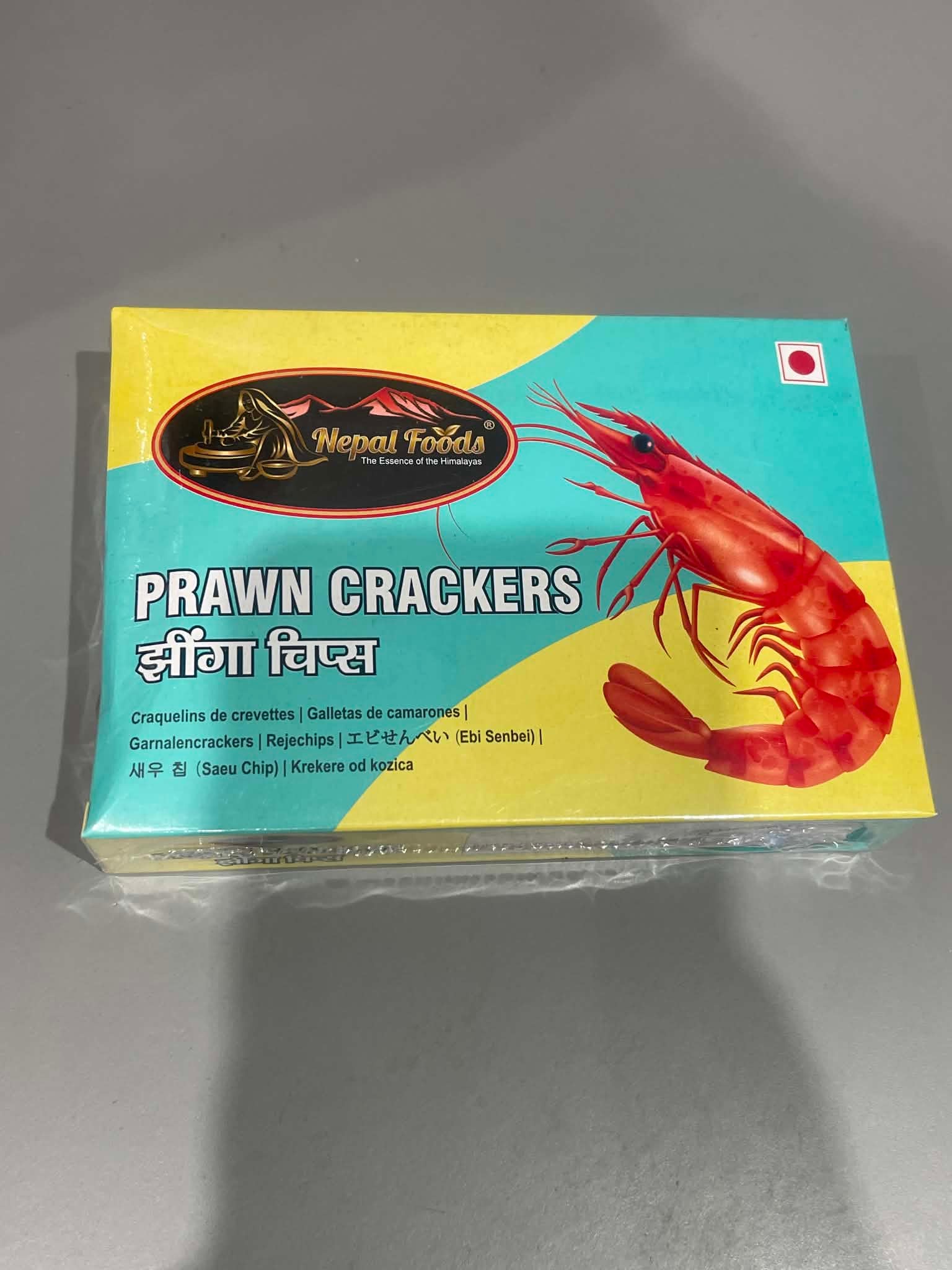 A boxed pack of Nepal Fable Prawn Crackers, featuring a bright turquoise and yellow design with a large red prawn illustration and a small Nepalese flag, photographed on a gray surface.