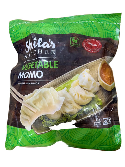 A package of Shila's Kitchen Vegetable Momo (wrinkle dumplings) on a light wood background. The packaging is green and dark brown, featuring a photo of the dumplings on a plate with dipping sauce.